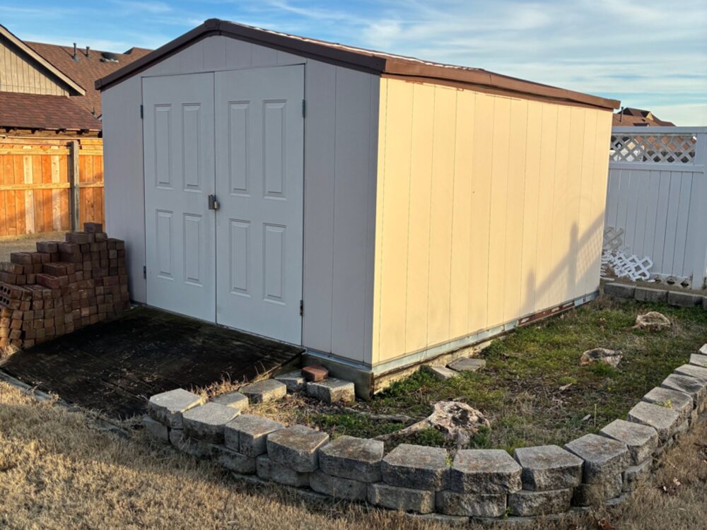 A garden shed standing on its slab beside a stacked pile of bricks and a stone border